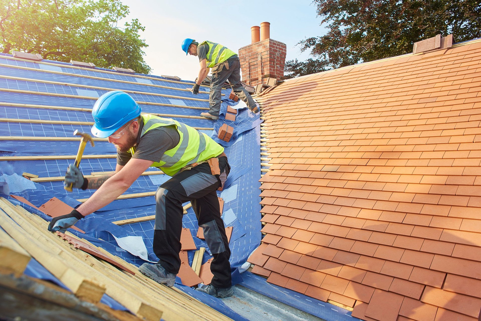 Construction workers in safety gear installing roofing materials on a residential house, with blue and terracotta colored roof panels visible.