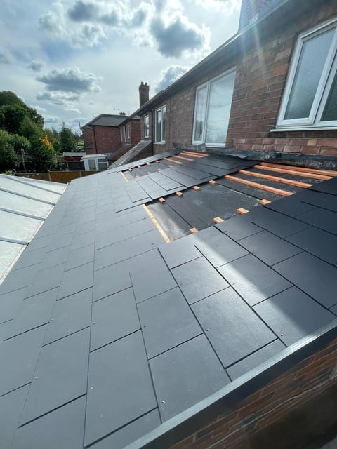 Roof installation in progress showing gray slate tiles on brick house with orange underlayment visible and sky in background