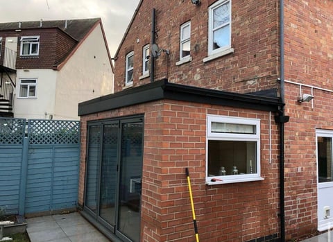 Red brick extension with black-framed glass doors and window, attached to residential home with blue gate visible on left